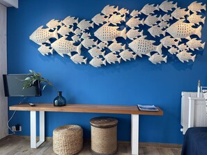 Ein Zimmer mit blauer Wand, einem Holztisch mit Vase und Fernbedienung, einem gewebten Hocker und einer Wanddekoration mit Fisch.