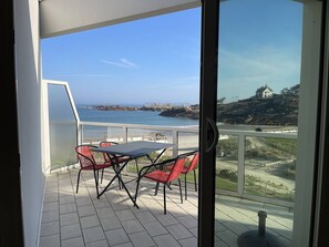 Ein Balkon mit Tisch und Stühlen, der Blick auf die Küste mit einem Haus auf einem Hügel freigibt.