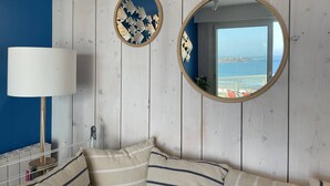 Quarto com cama, espelho redondo, abajur e vista para o mar através de uma janela.