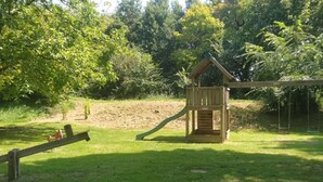Estructura de madera para juegos infantiles con tobogán y zona de escalada, rodeada de césped y árboles.