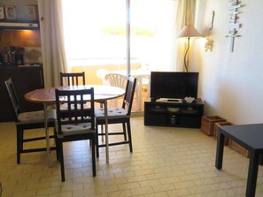 A dining area with a table and chairs, a television on a stand, and a lamp on the wall. - Seaside Apt with Parking, Beach Access (Sète)