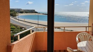 A balcony with a view of a beach and a road, featuring a table and chairs.