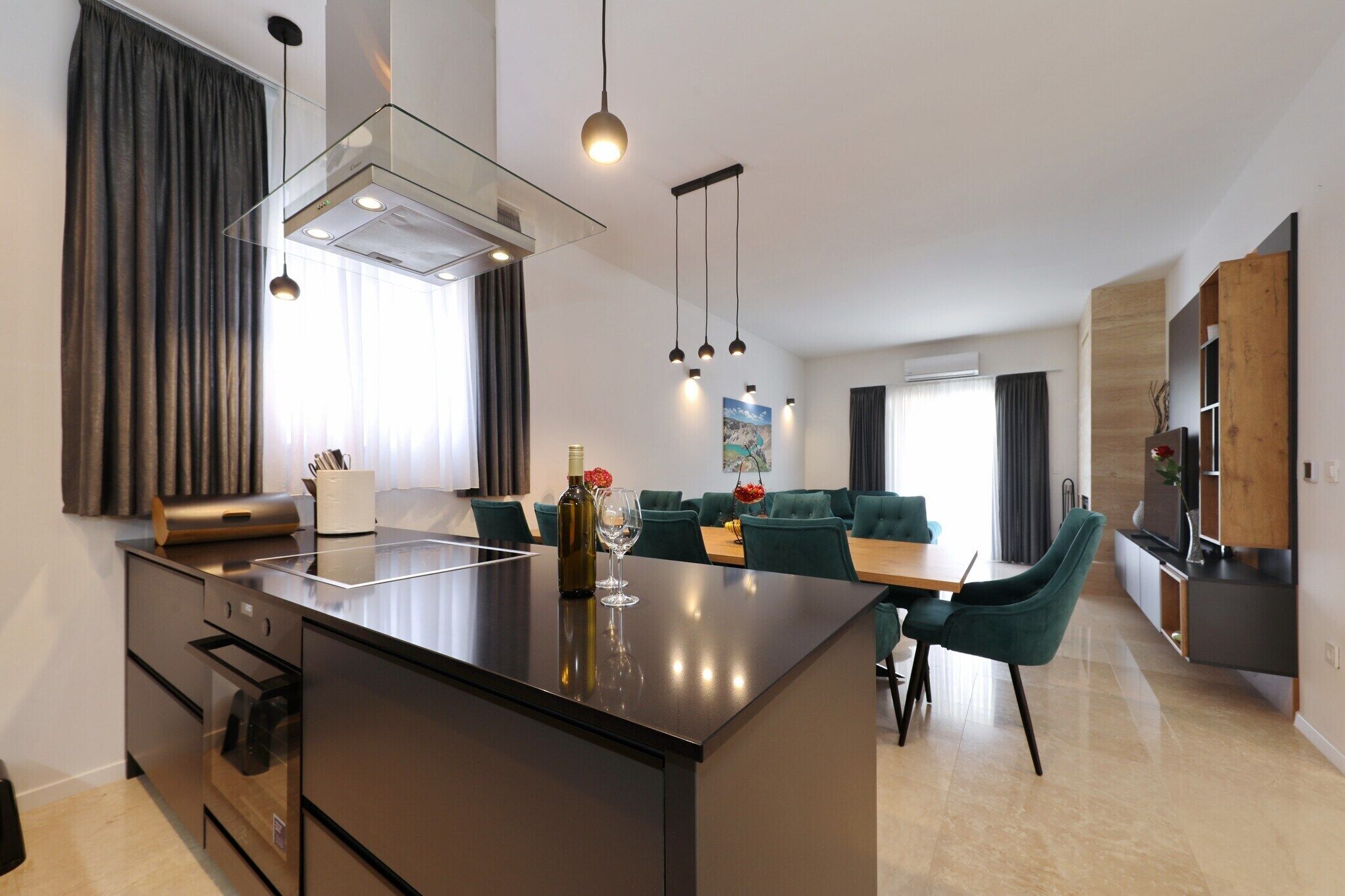 Una cocina moderna con una isla central, comedor con sillas verdes y un televisor a la derecha.