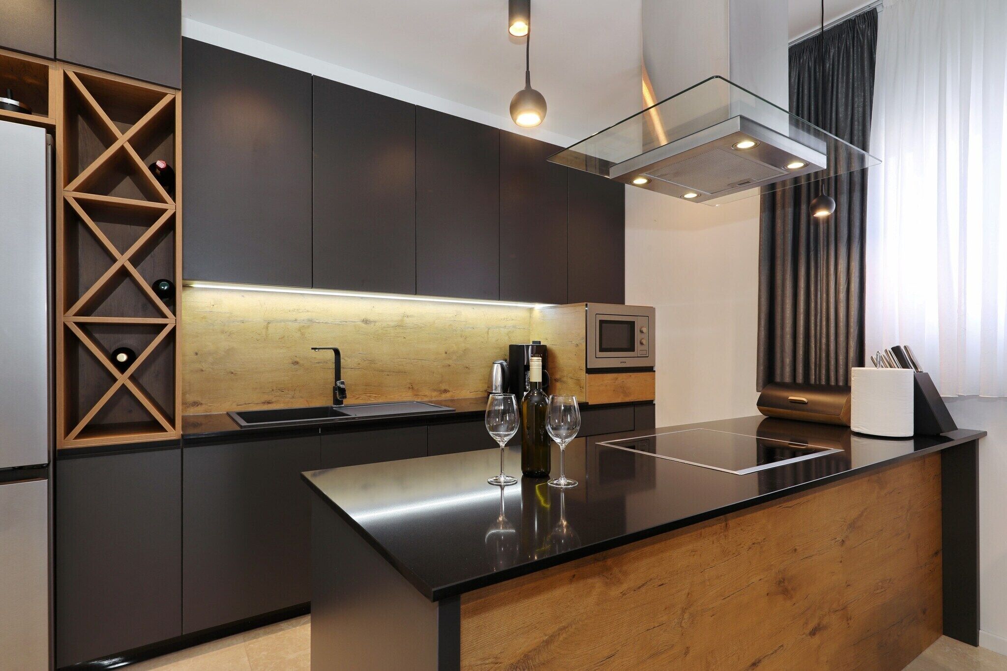 Une cuisine moderne avec des meubles de couleur sombre, un mur d’accent en bois, un îlot central en verre sur lequel sont disposés des verres à vin, et un four encastré.
