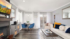 Ein modernes Wohnzimmer mit Kamin, einem Essbereich mit blauen Stühlen, einem weißen Sofa mit bunten Kissen und einem großen Flachbildfernseher an der Wand.