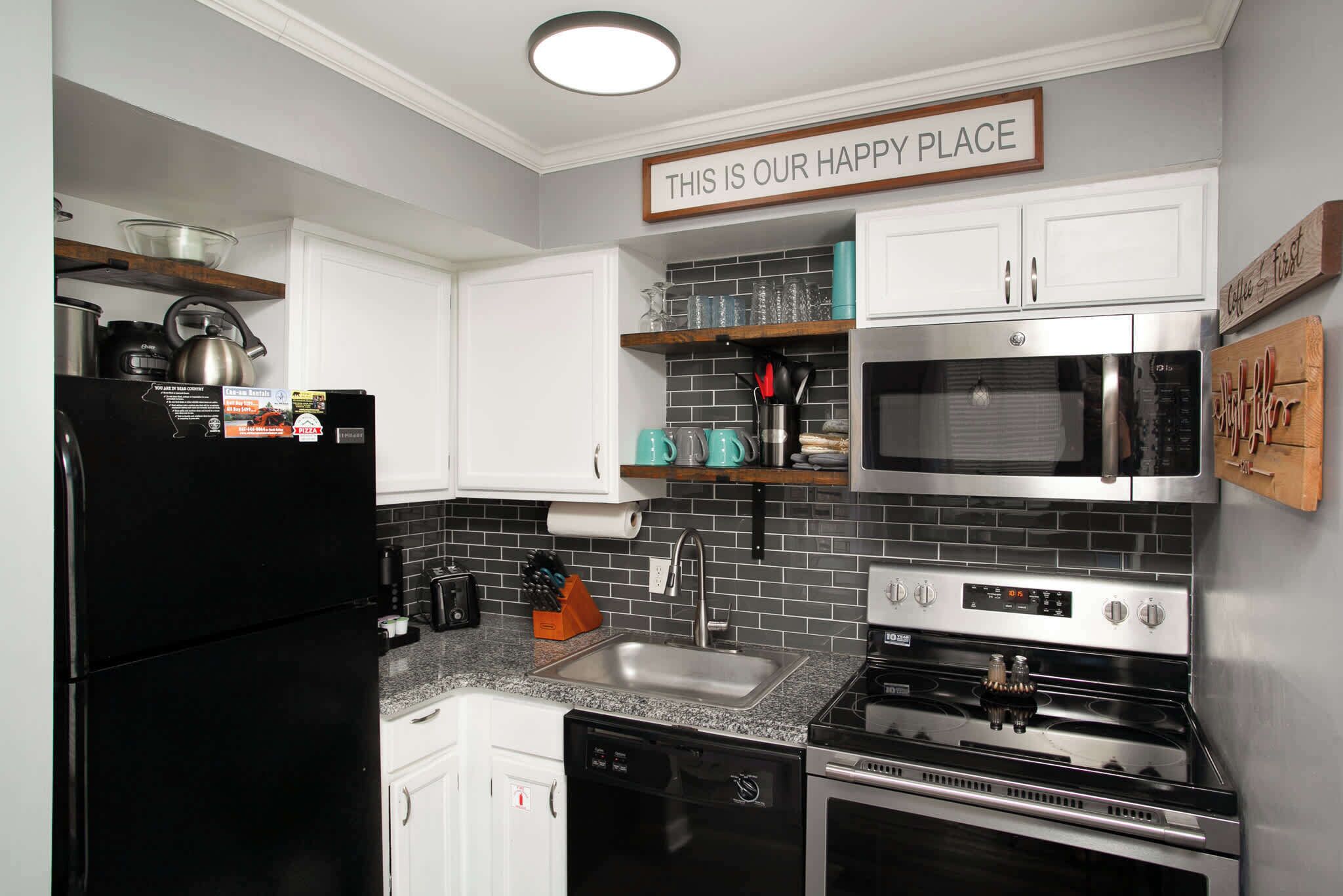 A modern kitchen with white cabinets, a black refrigerator, stainless steel appliances, and a sign that says "THIS IS OUR HAPPY PLACE".