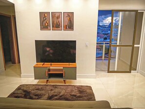 Een woonkamer met een flatscreen televisie, een houten tv-meubel en een groot raam met uitzicht op de stad.