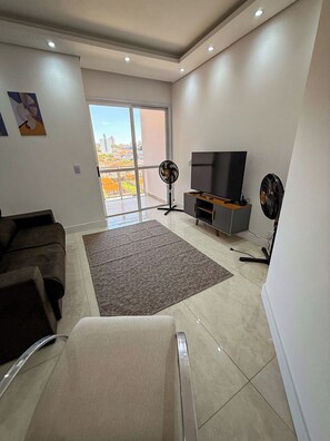 Een moderne woonkamer met een grote televisie, een bank, een ventilator en uitzicht op een stadsbeeld door een glazen deur.