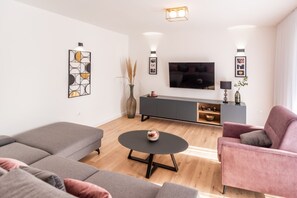 Una sala de estar moderna con un sofá gris, una mesa de centro redonda y un sillón rosa. Un televisor de pantalla plana está montado en la pared sobre un mueble gris.