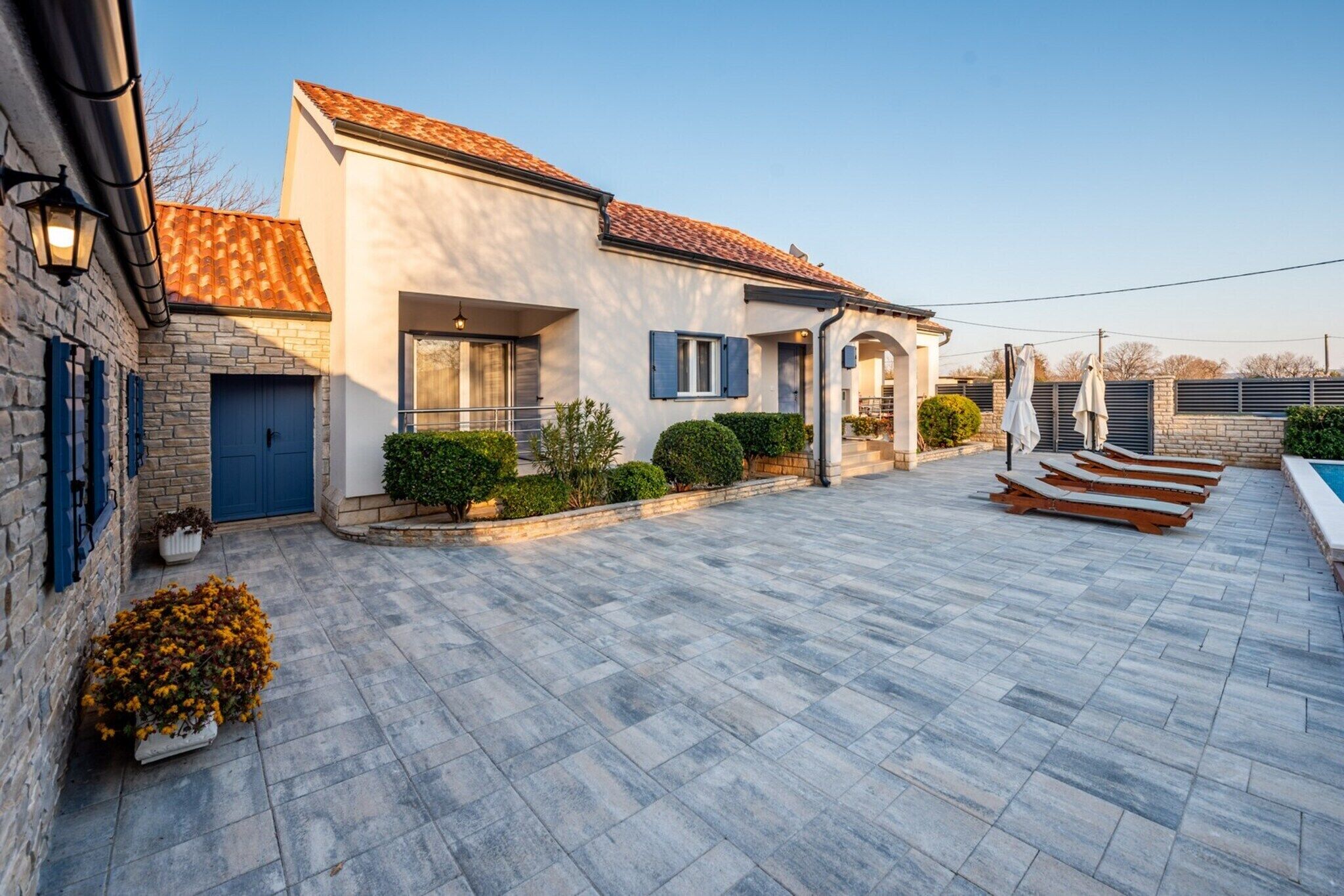 Une maison moderne avec un toit en tuiles, une façade en pierre et un patio dallé agrémenté de chaises longues.