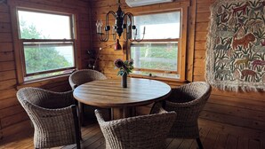 Interieur van een houten chalet met een ronde tafel, rieten stoelen, een kroonluchter en een wandtapijt met dierenmotieven.