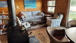 Een knus blokhutinterieur met een open haard, een bank, een rieten stoel en een schilderij aan de wand.