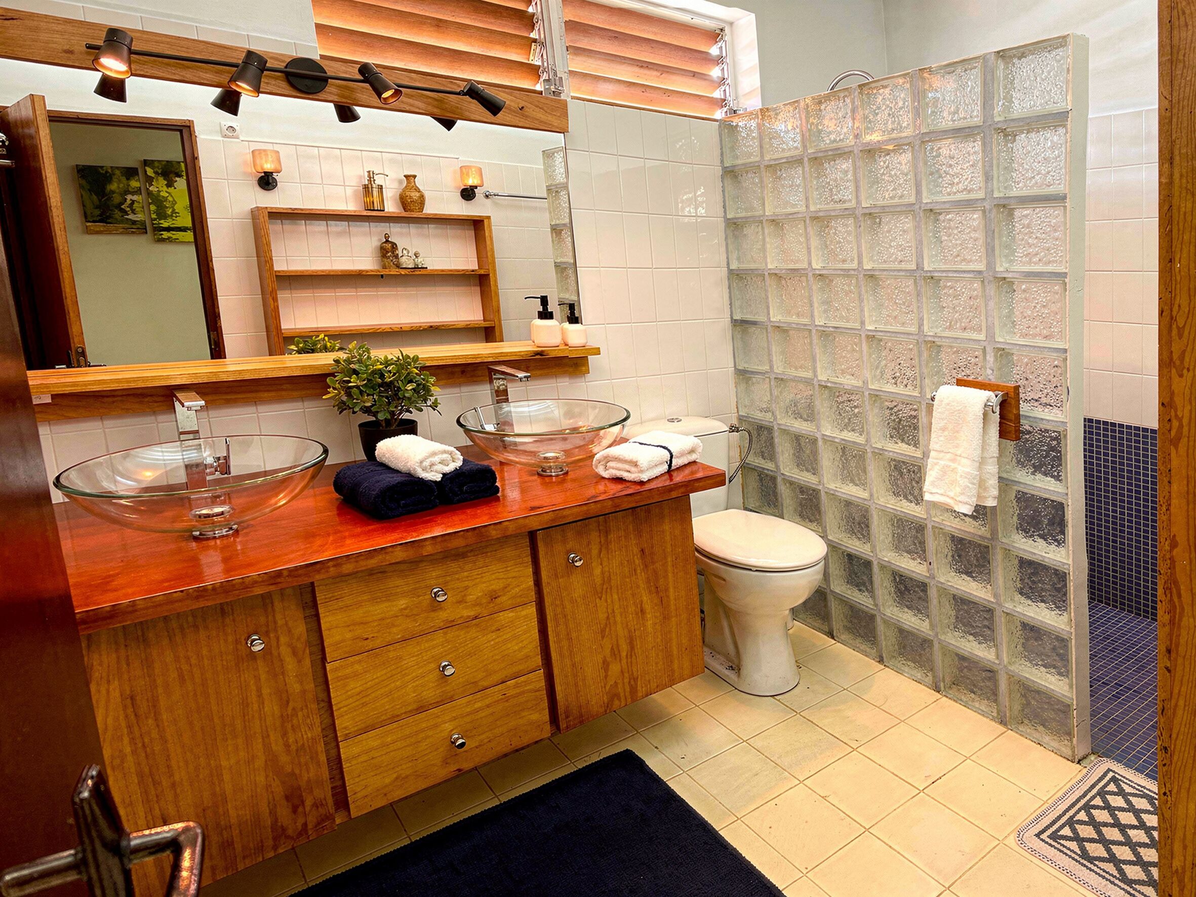 A bathroom with a wooden vanity, two glass bowls, a white toilet, and a frosted glass shower enclosure.