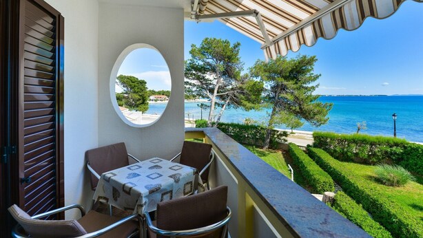 A balcony with a table and chairs, overlooking a clear blue sea and a well-maintained garden.