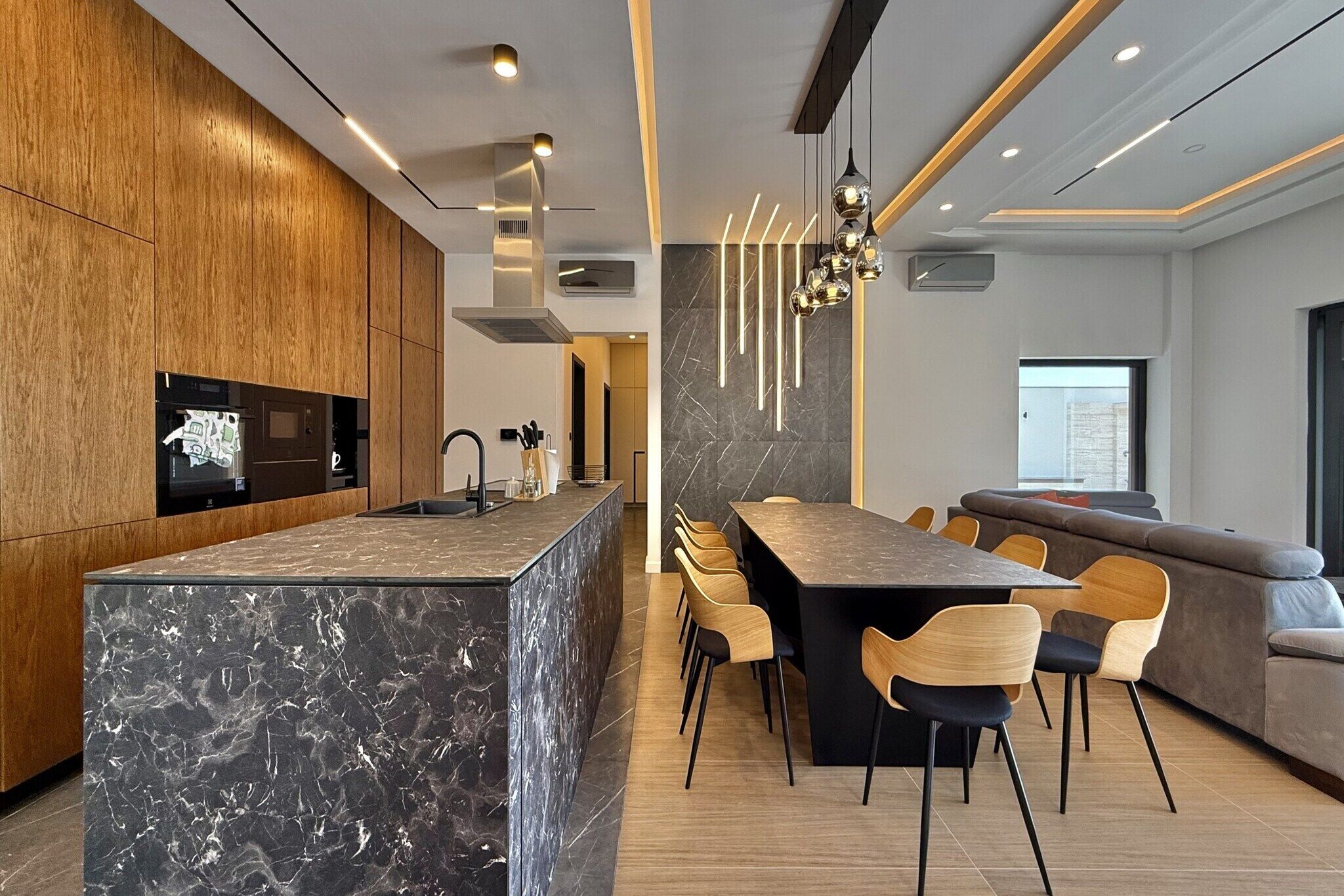 Une cuisine moderne dotée d’un îlot en marbre, de meubles en bois et d’un coin repas avec une longue table et des chaises.