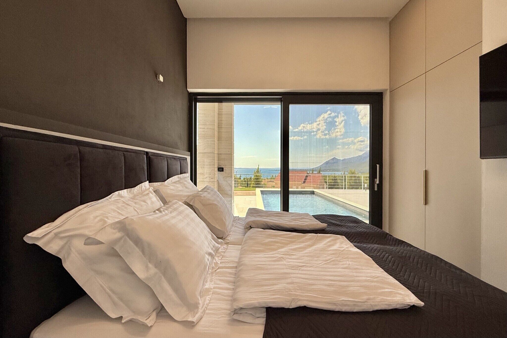 A modern bedroom with a large bed, a view of a swimming pool and mountains, and a sliding glass door to the outdoors.