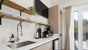 Cozinha moderna com lava-louças embutida, cafeteira, micro-ondas e pia. Prateleiras de madeira acima do balcão exibem louças e um vaso de planta.