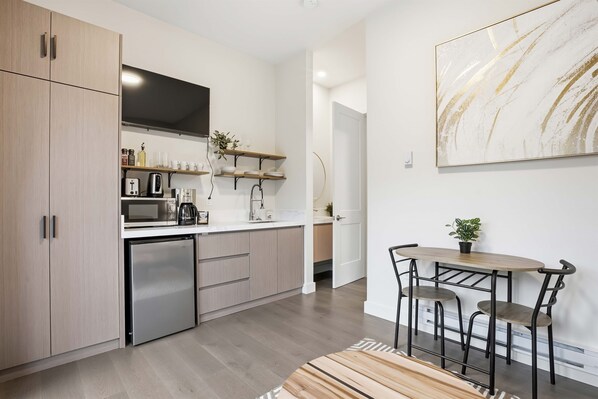 A modern kitchen with a built-in refrigerator, microwave, and coffee maker. There is a dining area with a table and chairs, and a potted plant on the wall. - Little Bear Den#2-Couples’ Cozy Minimalist Getaway (Ucluelet)