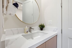 A bathroom with a white marble sink, a round mirror with a gold frame, a small potted plant, and a towel rack with a white towel and a dark towel. - Little Bear Den#2-Couples’ Cozy Minimalist Getaway (Ucluelet)