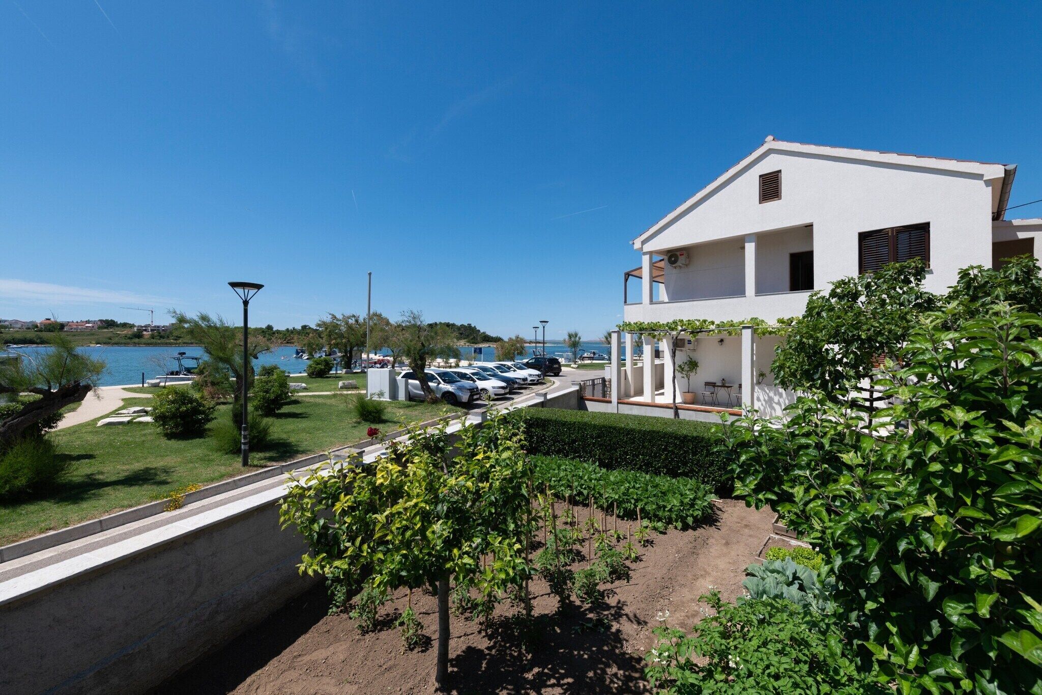 A modern white building with a balcony, a parking area with several cars, and a view of the sea.