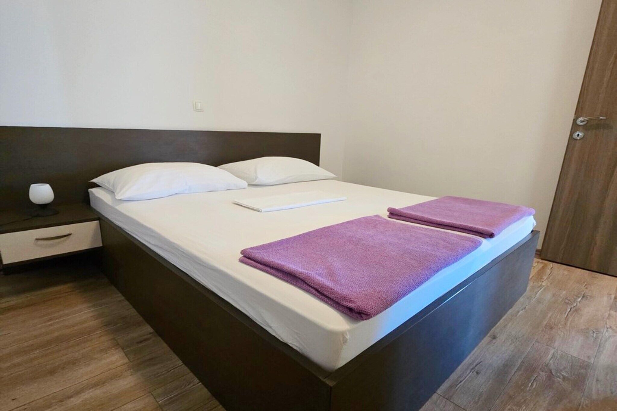 A neatly made bed with white linens and two purple towels, a bedside table with a lamp, and a wooden floor.