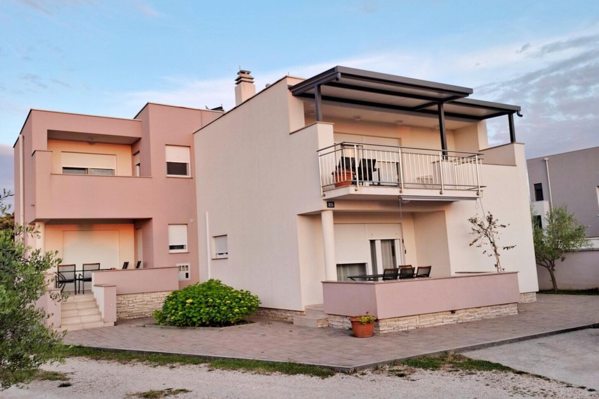 A modern two-story residential building with a balcony, a small garden, and a paved driveway.