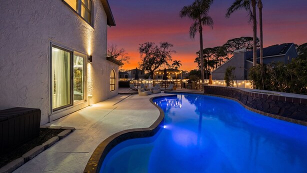 Ein Pool mit geschwungener Kante, eine Terrassenanlage mit Gartenmöbeln und ein Haus mit großen Fenstern und einer Steinmauer.