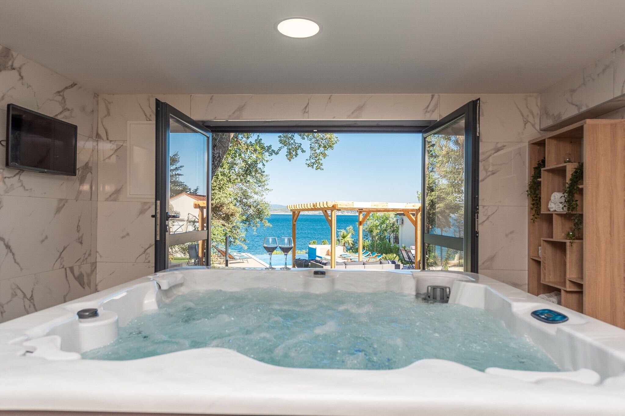 Une salle de bain moderne avec une grande baignoire, un téléviseur fixé au mur et une vue sur la mer par une porte ouverte.