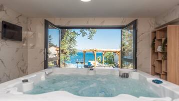 Une salle de bain moderne avec une grande baignoire, un téléviseur fixé au mur et une vue sur la mer par une porte ouverte.