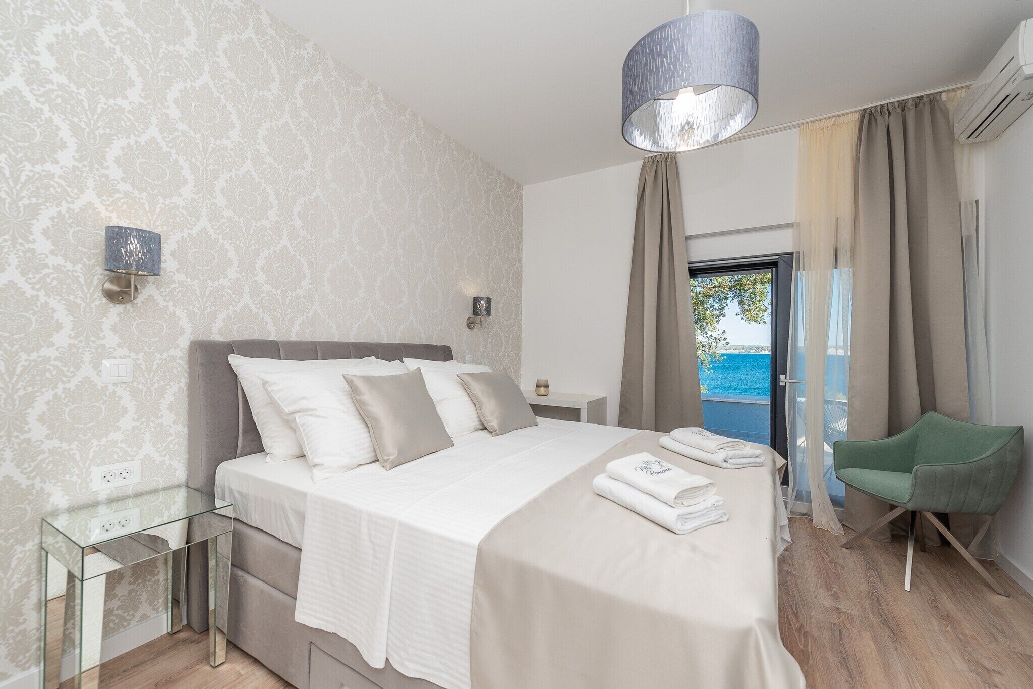 Une chambre à coucher moderne avec un lit, des tables de chevet, un fauteuil vert et une vue sur la mer.