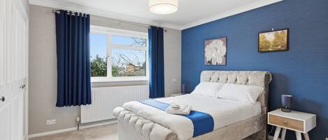 A bedroom with a large bed, a bedside table, a window with curtains, and a blue accent wall.