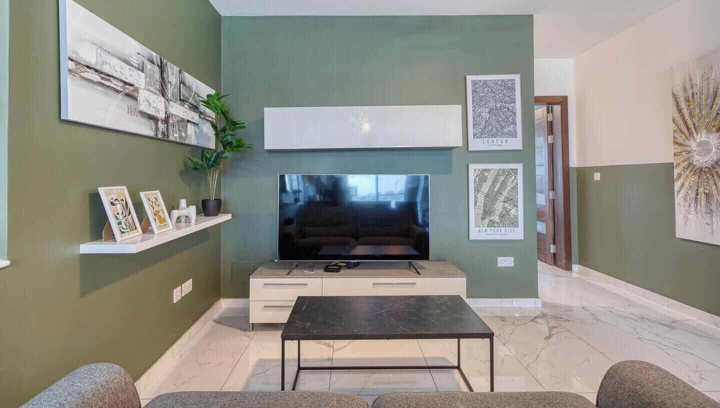 A living room with a flat-screen TV, a coffee table, and a sofa. There are framed pictures on the wall and a plant on a shelf.