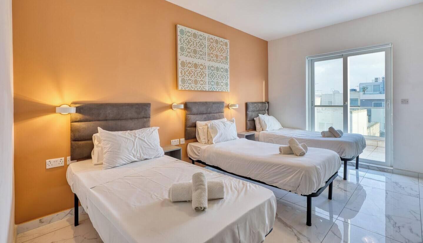Una camera d'albergo con tre letti, ognuno dotato di testiera e lampade sui comodini. Ampia finestra con vista sulla città.