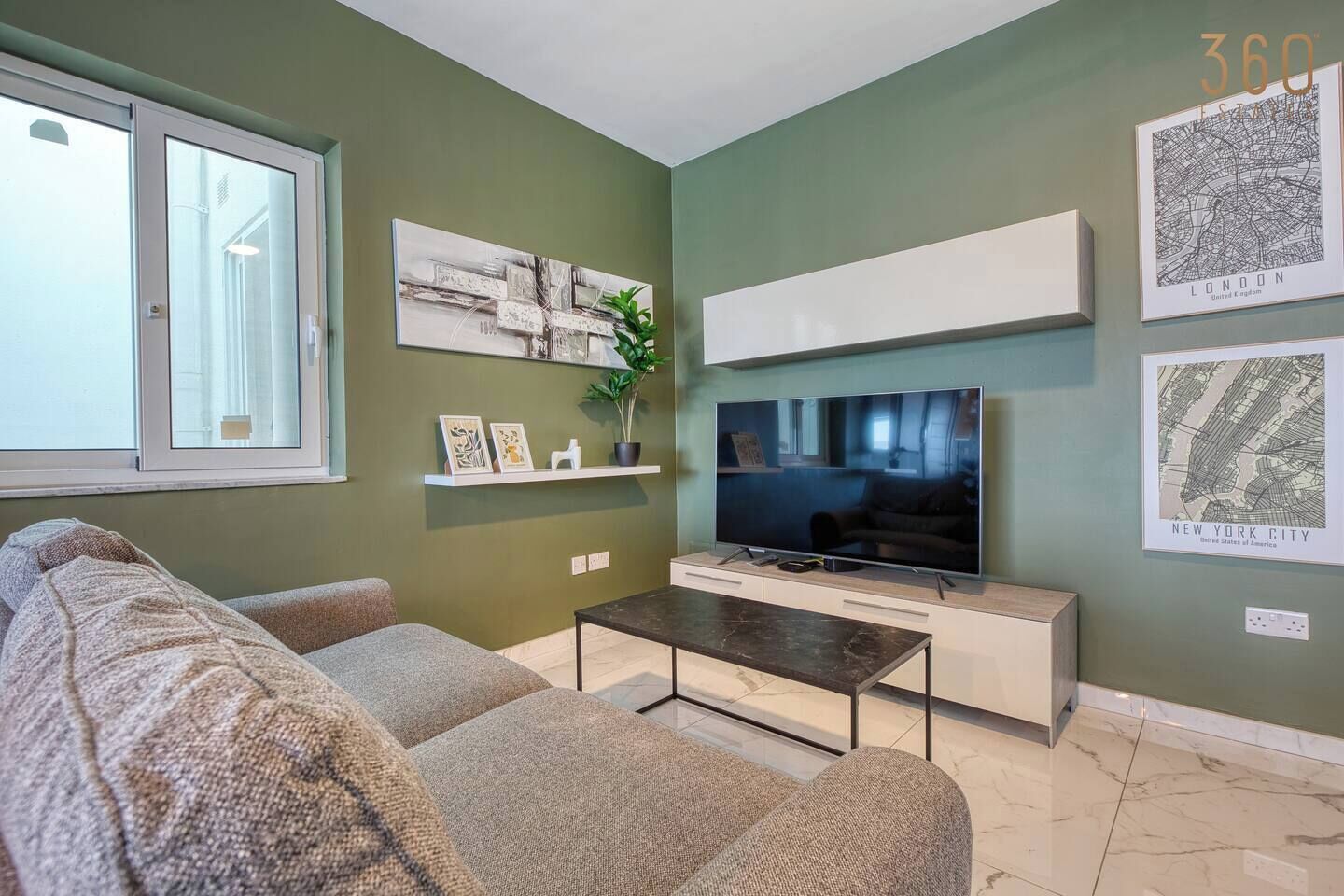 A living room with a sofa, a coffee table, a flat-screen TV, and framed city maps on the wall.