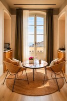 Una habitación con una mesa redonda, dos sillas de mimbre, un ventanal con cortinas y vistas a edificios.