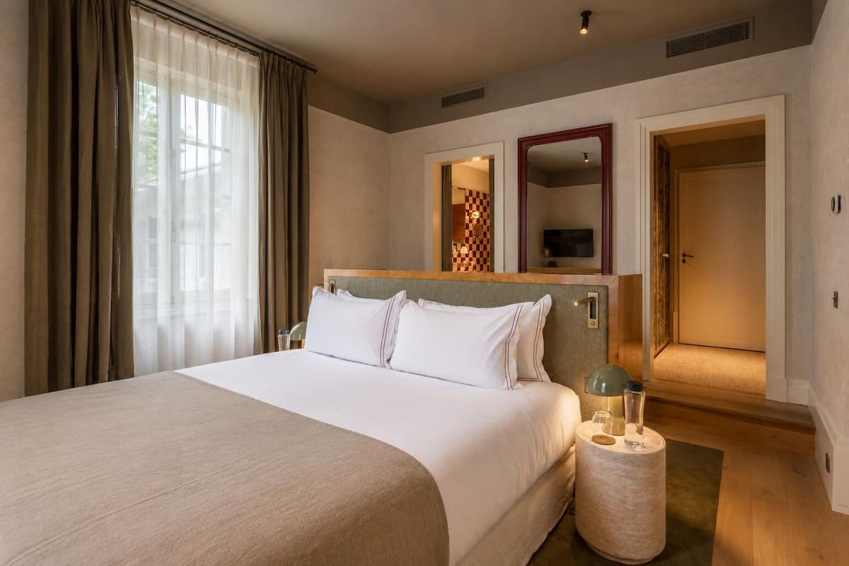 A hotel room with a large bed, a bedside table with a lamp, a mirror, and a door leading to another room.