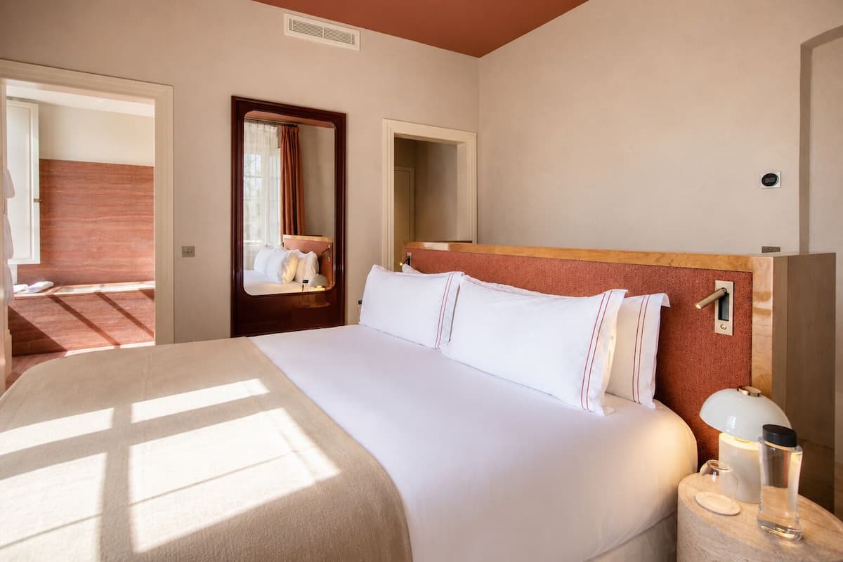 A hotel room with a large bed, a bedside table with a lamp, a mirror, and a door leading to another room.