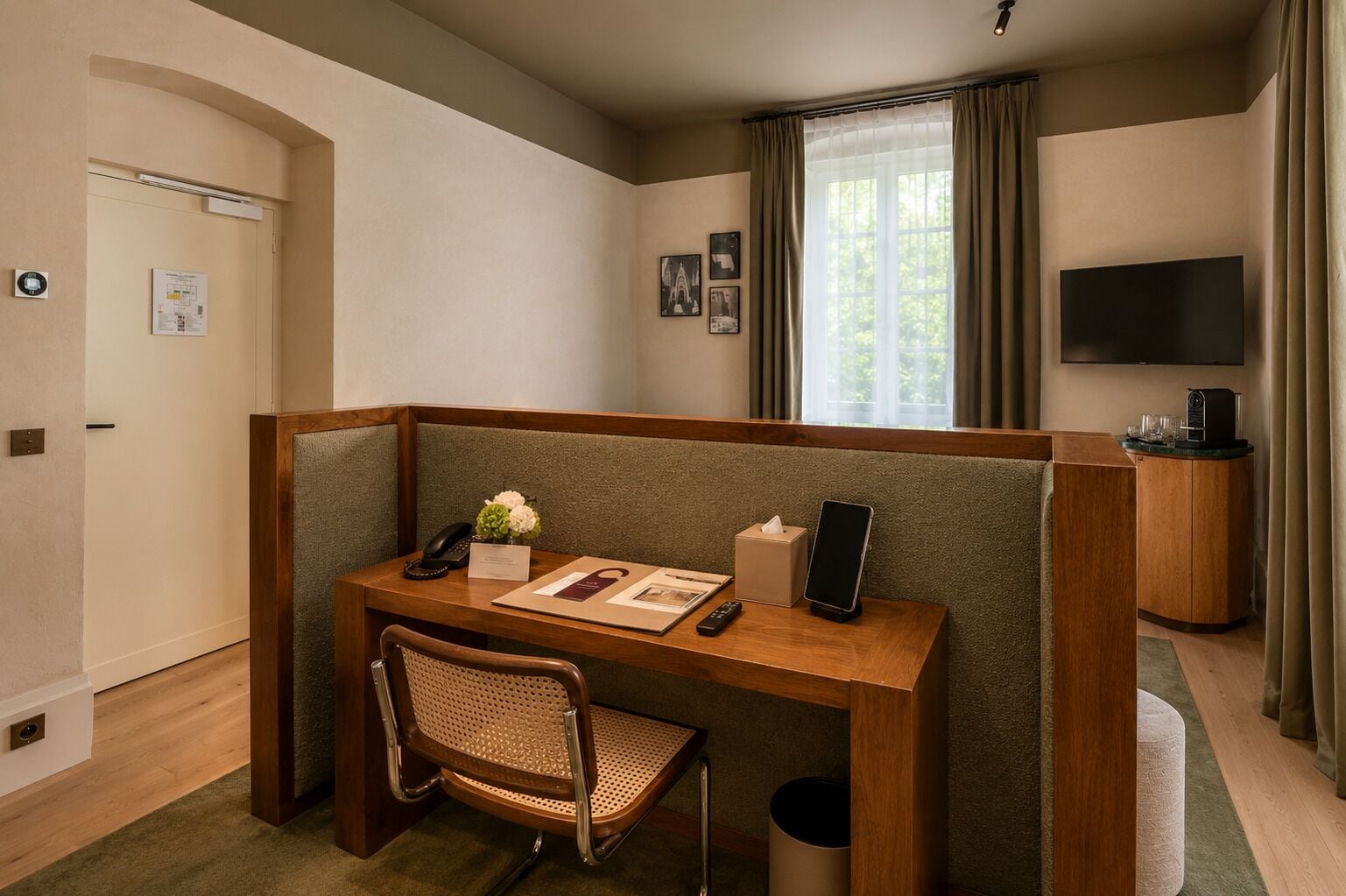 Une chambre d’hôtel avec un bureau en bois, une chaise, un téléphone, une boîte de mouchoirs, une télécommande, une tablette, un téléviseur et une fenêtre avec des rideaux.