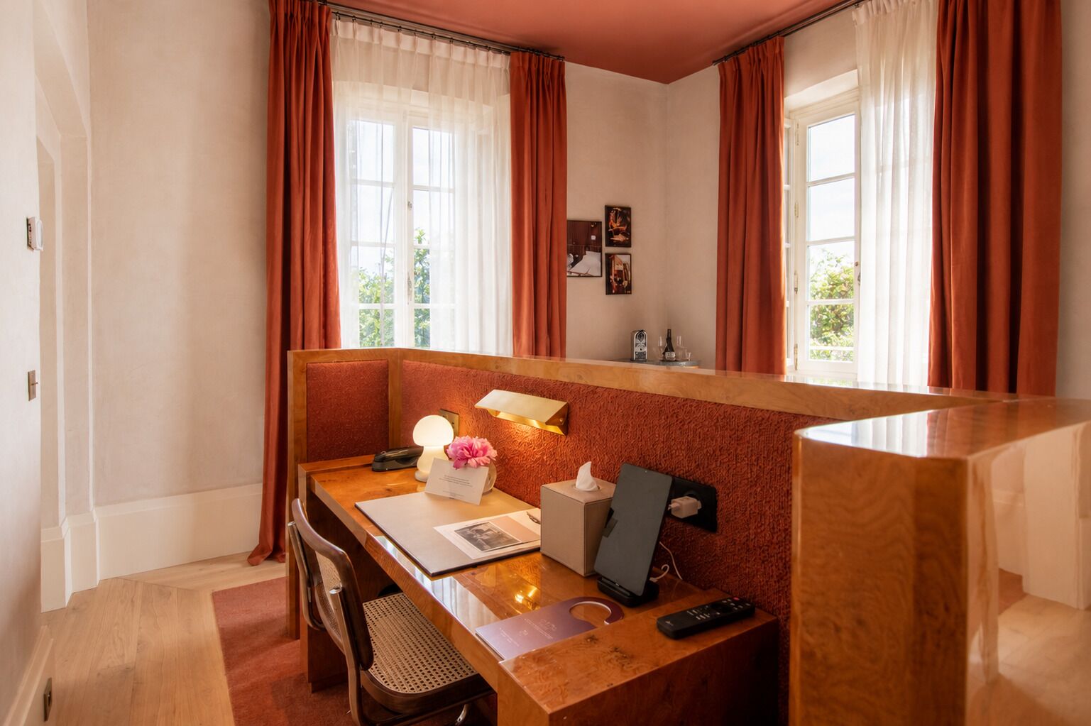 A hotel room with a wooden desk, a chair, a bedside table, a lamp, a remote control, and a window with curtains.