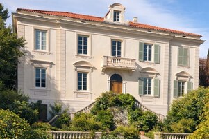 Un edificio grande de dos pisos con fachada simétrica, múltiples ventanas con contraventanas y una entrada central con puerta.