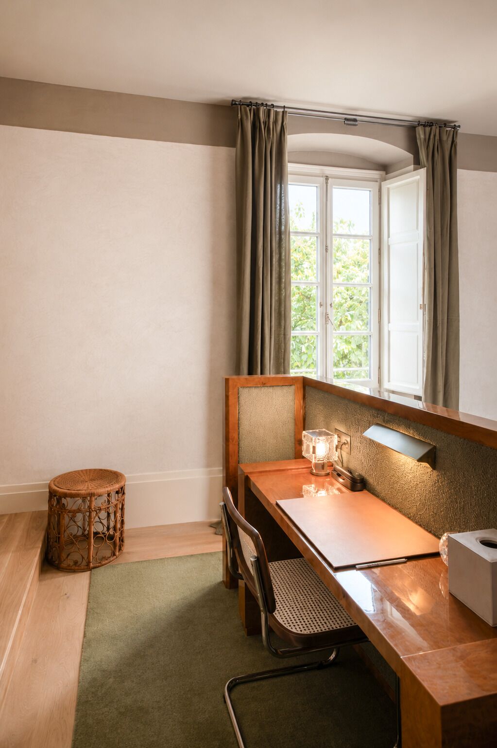 Una habitación con un escritorio de madera, una silla, un taburete redondo de mimbre, una lámpara y una ventana con cortinas.