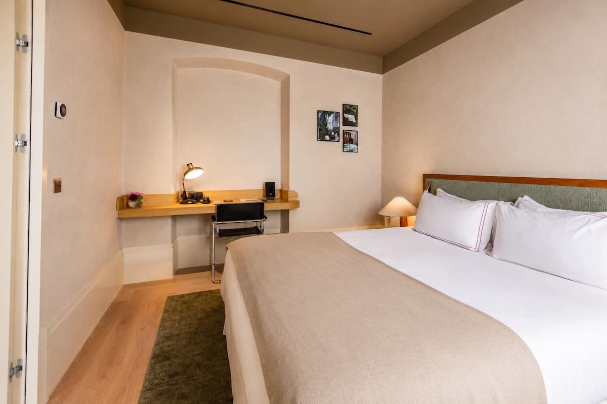A hotel room with a bed, a desk with a lamp, a chair, a small table with flowers, and a picture on the wall.