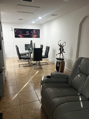 A modern living room with a leather sofa, a dining area with a glass table and chairs, and a mounted artwork above the table.