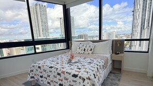 Un dormitorio moderno con una cama grande, una mesita de noche y vistas de edificios altos a través de amplias ventanas.