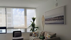 Un salón moderno con un sillón gris, un sofá beige con cojines estampados, un ventanal con cortinas translúcidas, un cuadro de un paisaje urbano y una pequeña mesa redonda con un letrero que dice 'WELCOME'.