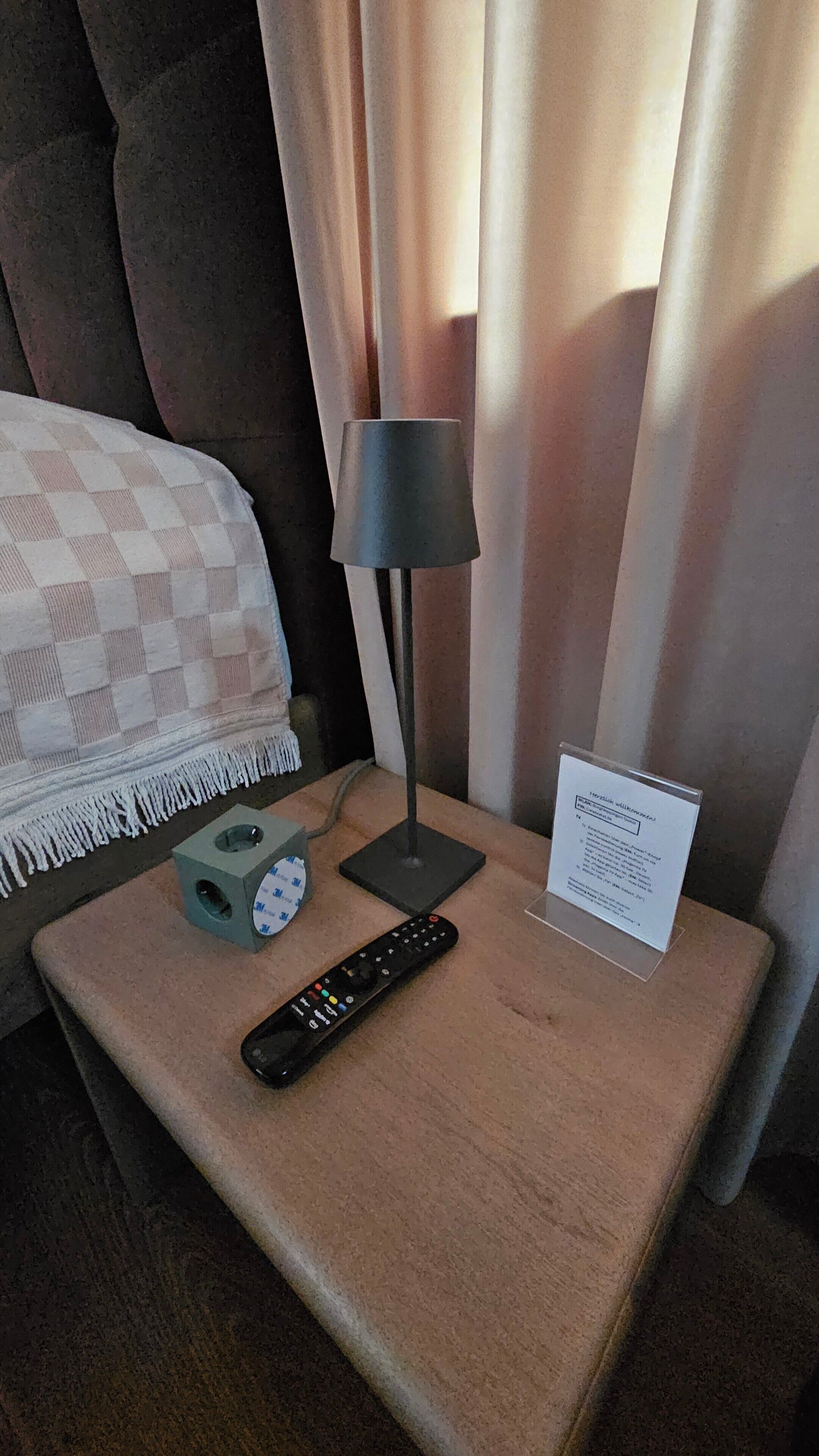 Habitación de hotel con cama, mesita de noche, lámpara, control remoto y cartel.