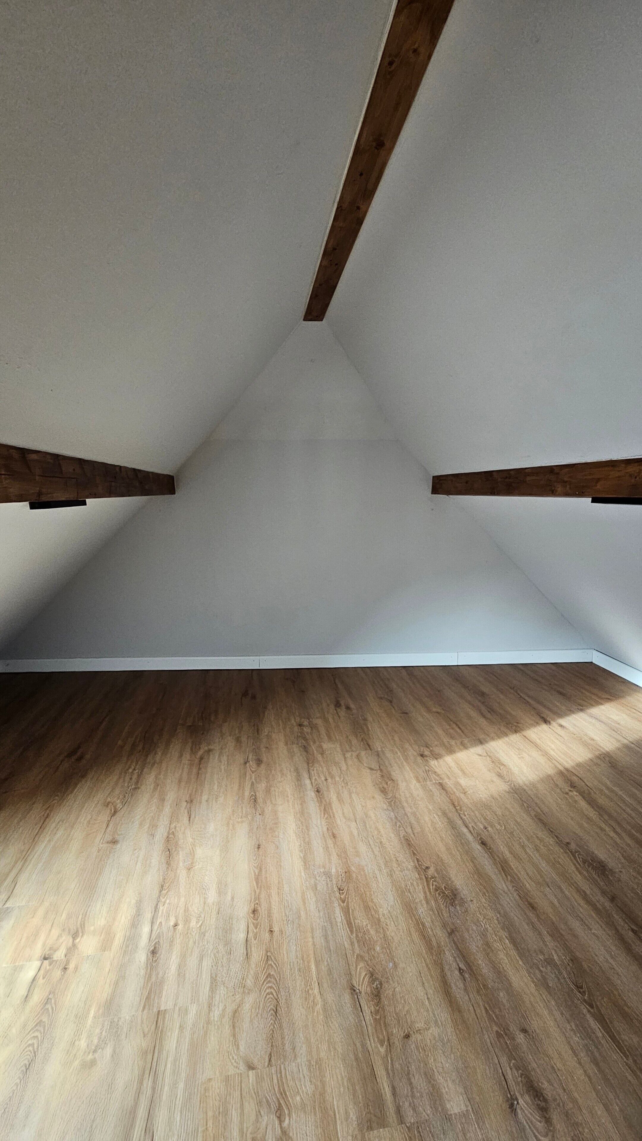 Una stanza mansardata con travi in legno, soffitto spiovente e pavimento in legno.