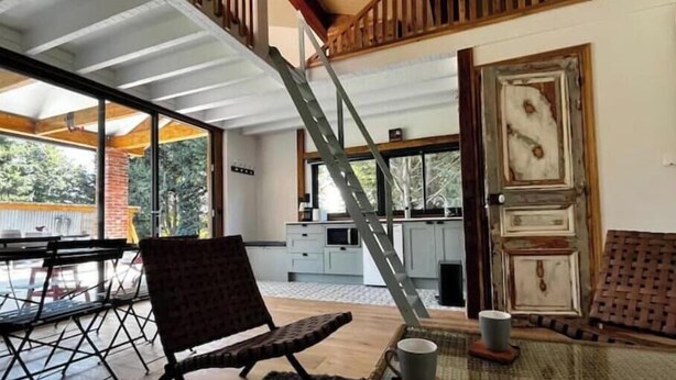 Una habitación amplia con techo alto, un comedor con mesa y sillas, una zona de cocina con armarios, y un gran ventanal que ofrece vistas al exterior.