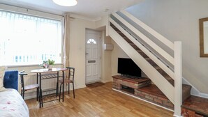 A room with a wooden floor, a dining table with chairs, a TV on a brick fireplace, a staircase with white railings, and a window with curtains. - Headmasters Hollow (Watford)
