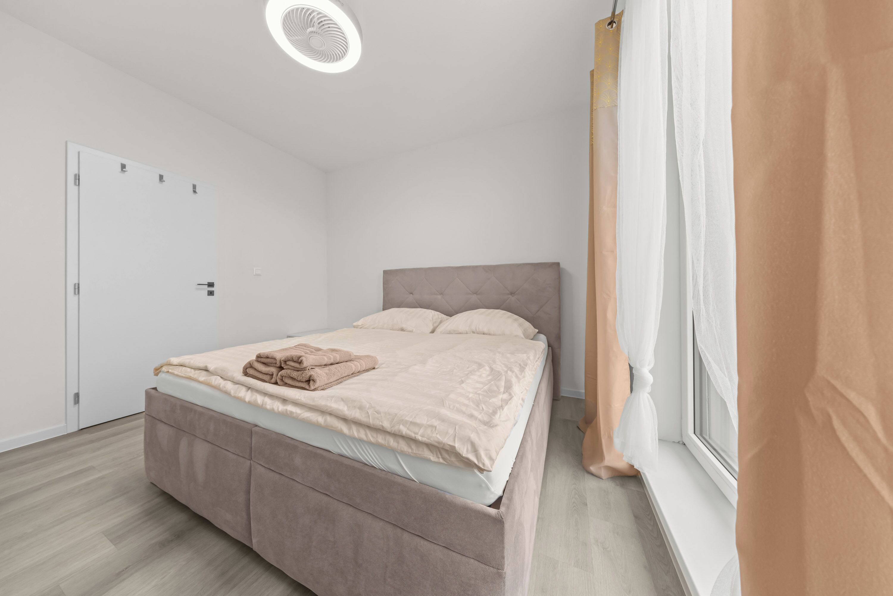 Een slaapkamer met een bed, een raam met gordijnen, een plafondventilator en een deur.